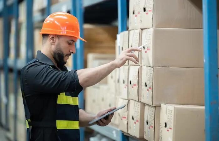 Trabajador en almacén con casco inspecciona cajas usando una tableta, ilustrando la gestión de stocks