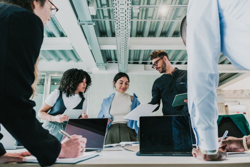 On voit une équipe (deux hommes de dos, deux femmes et un homme de face) en train de travailler ensemble autour d'un logiciel de gestion du parc informatique, sur leurs ordinateurs