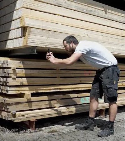 Trabajador escaneando el código de barras de pilas de madera con su móvil para registrar materiales en el almacén.