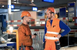 Dos trabajadores de la construcción con chaleco y casco, hablando en la obra de la estación de tren de Berna.
