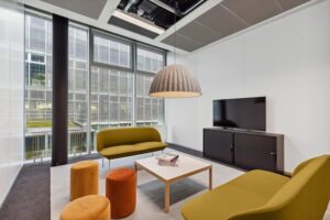 ista de una sala de reuniones moderna y luminosa en FlexOffice, con muebles coloridos y grandes ventanales.