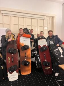 Equipo de Salty Lemon sonríe y sostiene tablas de kitesurf en el almacén, rodeados por equipamiento preparado para el alquiler.