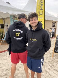 Dos miembros del equipo Salty Lemon, vestidos con sudaderas negras y pantalones deportivos, conversan en la playa durante una jornada de kitesurf.