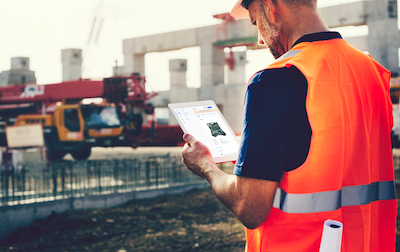 Trabajador de la construcción con chaleco reflectante usando una tableta en la obra para gestionar equipos con software de inventarios