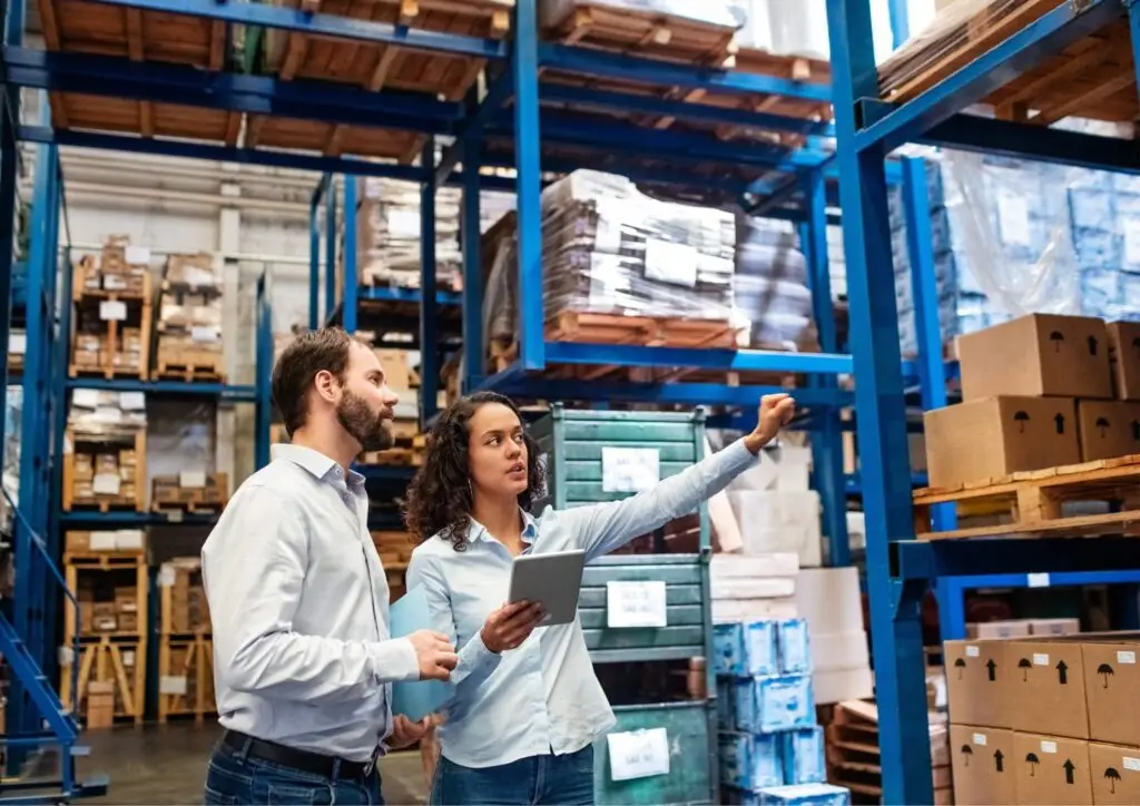 Dos trabajadores revisando el inventario de un almacén con ayuda de una tablet entre estanterías de cajas y palets.