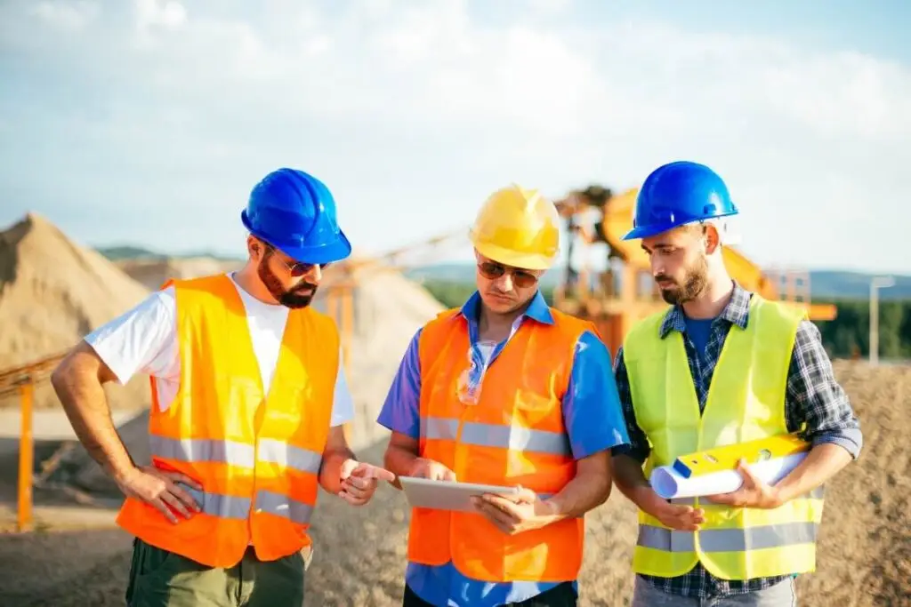 Trois ouvriers en casque et gilet haute visibilité consultent une tablette sur un chantier de carrière, illustrant la gestion des chantiers dans le BTP.