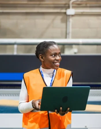 Trabajadora de mantenimiento en una nave industrial revisa tickets digitales en su portátil, sustituyendo el papeleo por un sistema de tickets con notificaciones automáticas y documentación jurídicamente segura